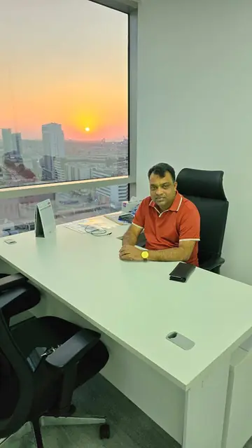 Abdul Kader Chowdhury at an office desk by the window at sunset, with the city skyline behind
