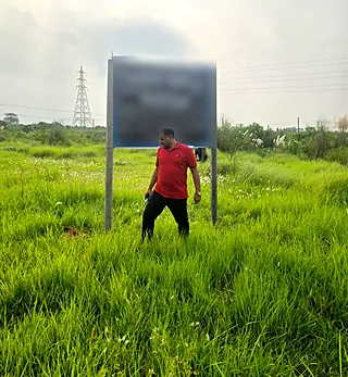 A visitor in a red shirt on green land in front of a land signboard (signboard text blurred)
